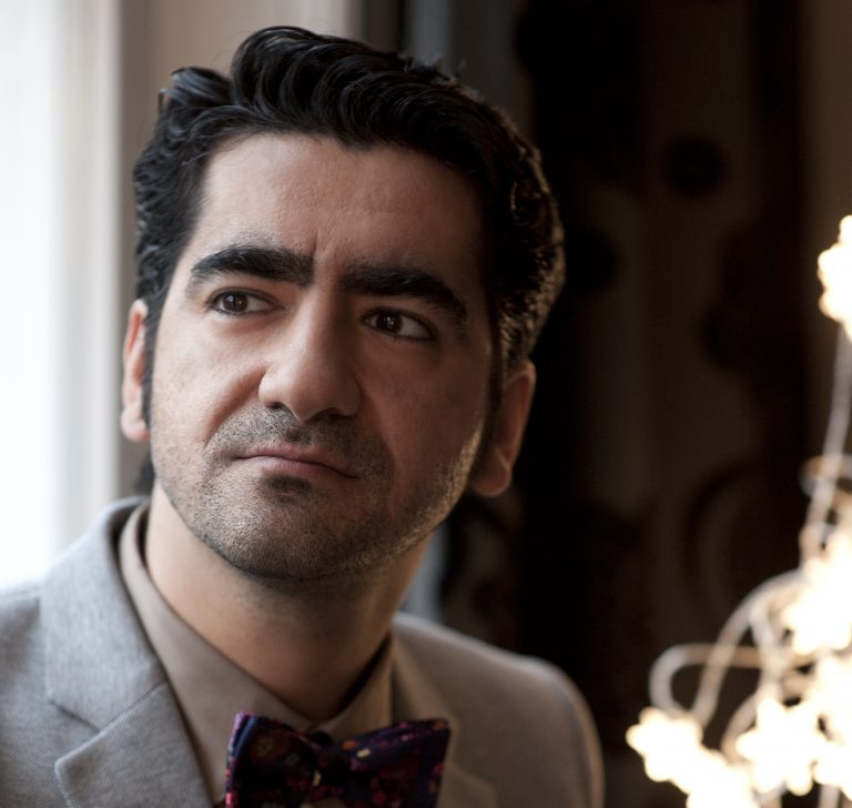Een man met een lichte grijze blazer en een gekleurde vlinderdas, die nadenkend naar buiten kijkt.