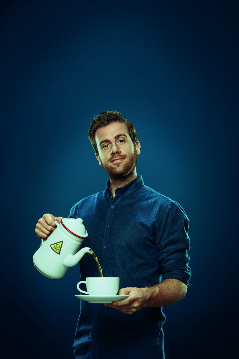 Een man in een blauwe shirt giet koffie uit een witte ketel in een kopje tegen een donkerblauwe achtergrond.