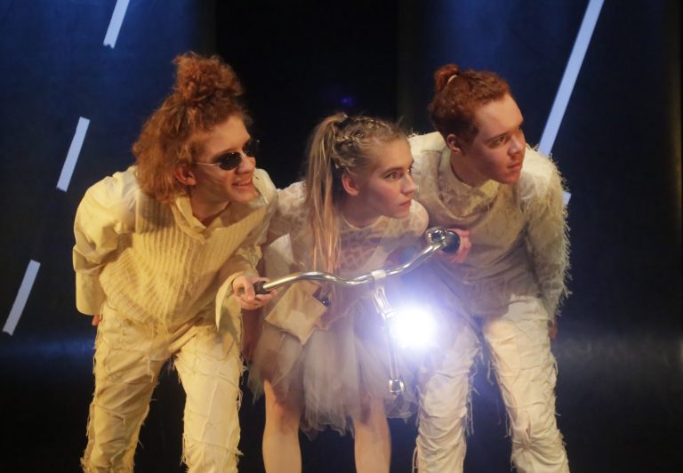 Drie acteurs in opvallende kostuums staan samen op een podium met een fiets, terwijl ze in de richting van de kijker kijken.