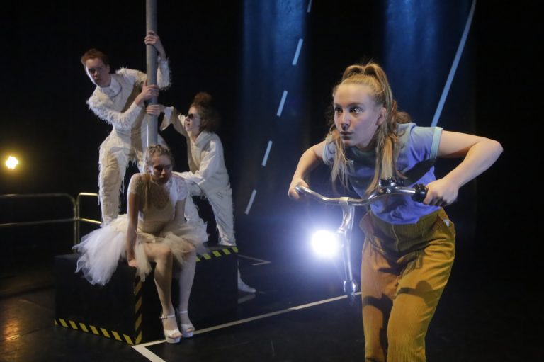 Een actrice op een fiets in een theaterproductie, omringd door andere performers in kostuums.