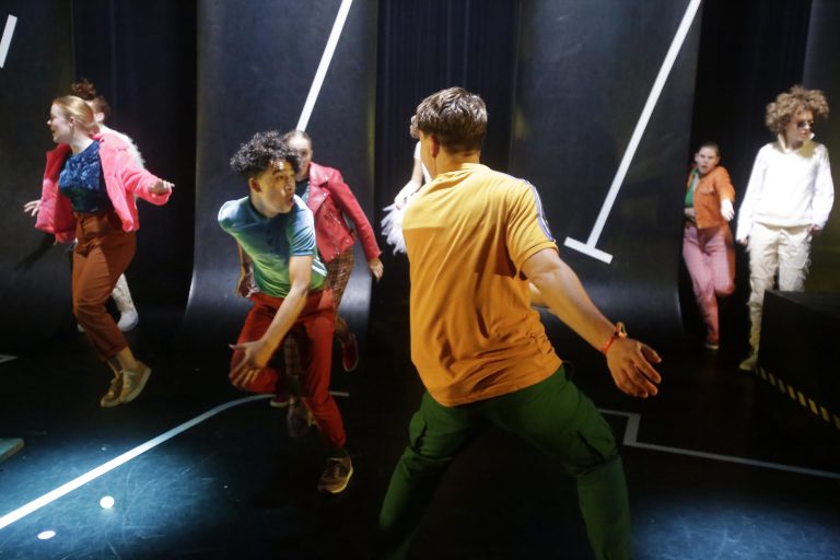 Groep dansers in kleurrijke outfits voert een dynamische choreografie uit op een donkere achtergrond.