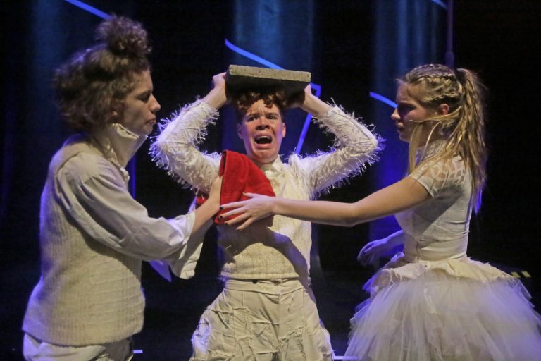 Drie acteurs zijn op het podium aan het spelen, waarbij één actievoerende persoon lijkt te worden vastgehouden en een boek op zijn hoofd heeft.