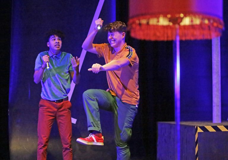 Twee performers in kleurrijke kleding voeren een energieke act op een podium uit.