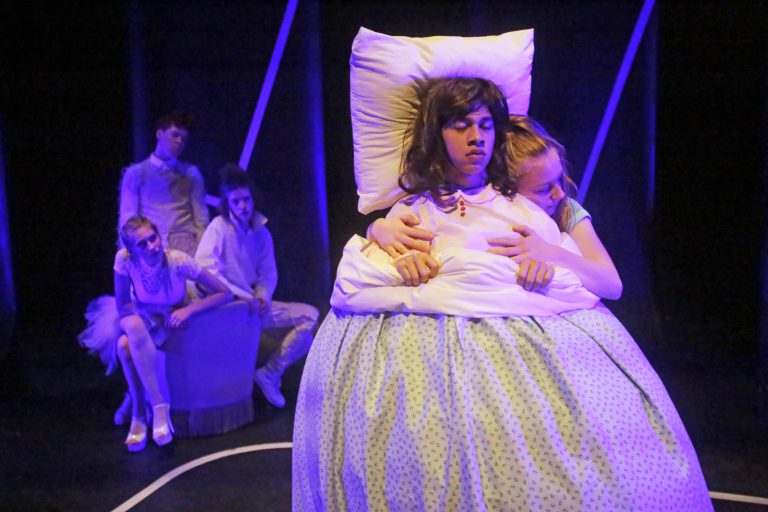 The image features a theatrical performance with a central character in bed, surrounded by other performers in a dimly lit setting.