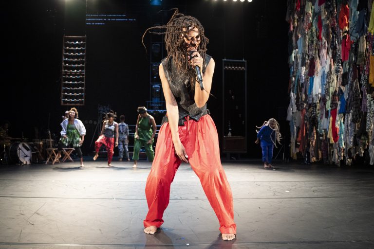 Een performer met dreadlocks staat op een podium en zingt in een flamboyante outfit met rode broek terwijl andere artiesten op de achtergrond te zien zijn.
