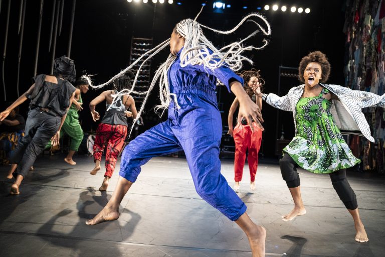Een groep dansers voert energieke bewegingen uit op een podium, met krachtige, kleurrijke kostuums en een dynamische uitstraling.