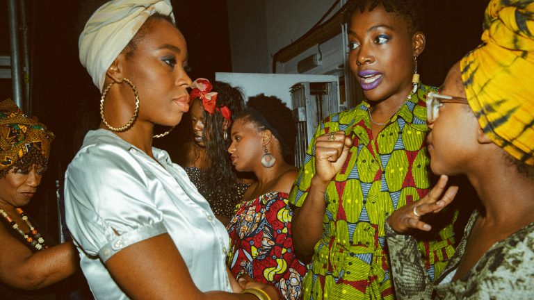 Twee vrouwen in levendige kleding voeren een gesprek, terwijl anderen op de achtergrond staan.