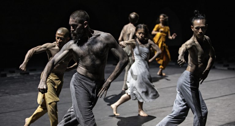Een groep dansers in verschillende kostuums voert een expressieve choreografie uit op een donkere achtergrond.