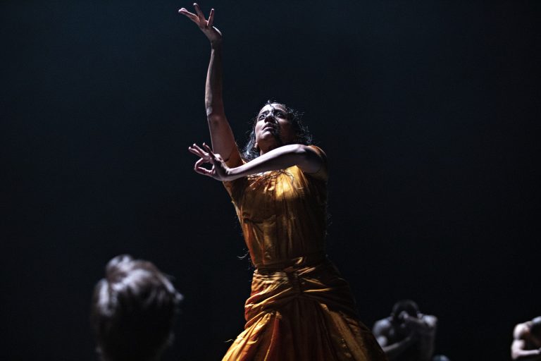 Een danseres in een gouden jurk voert een expressieve beweging uit op een donkere achtergrond.