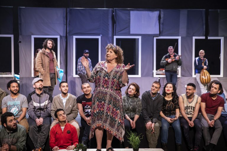 Een groep acteurs op een theaterpodium, met een centraal figuur die enthousiast een verhaal vertelt terwijl anderen toekijken.