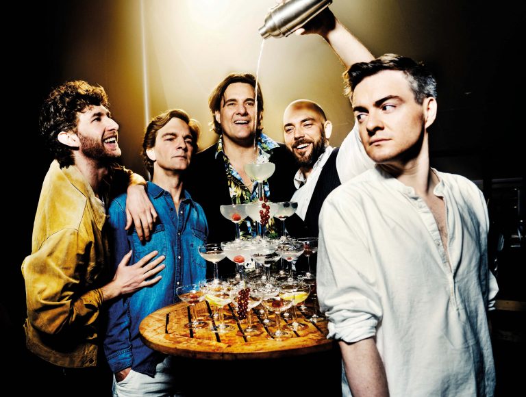 Een groep mannen poseert met een dienblad vol champagneglazen, terwijl ze vrolijk met elkaar interactie hebben.