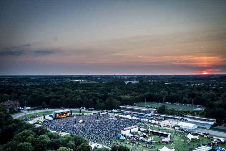Een luchtfoto van een groot festivalterrein met een druk publiek en een kleurrijke zonsondergang op de achtergrond.