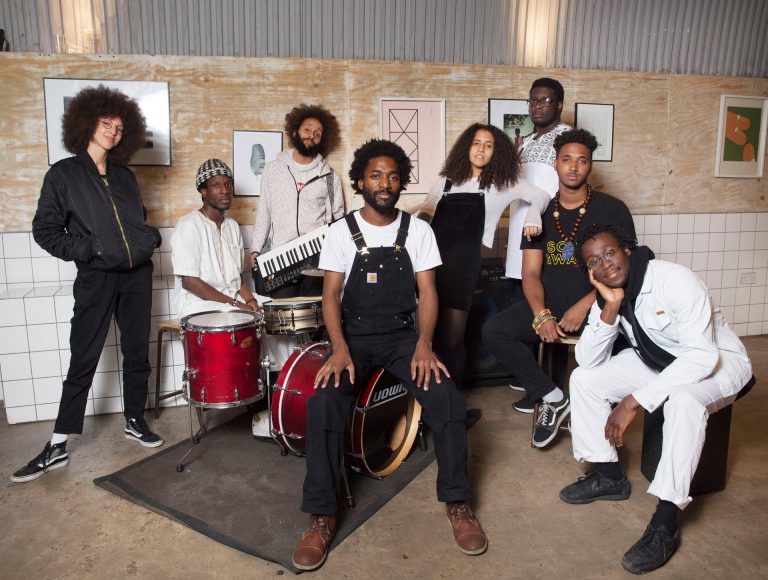 Een diverse groep van acht muzikanten poseert samen in een industriële ruimte, met verschillende muziekinstrumenten en informele kledingstijlen.