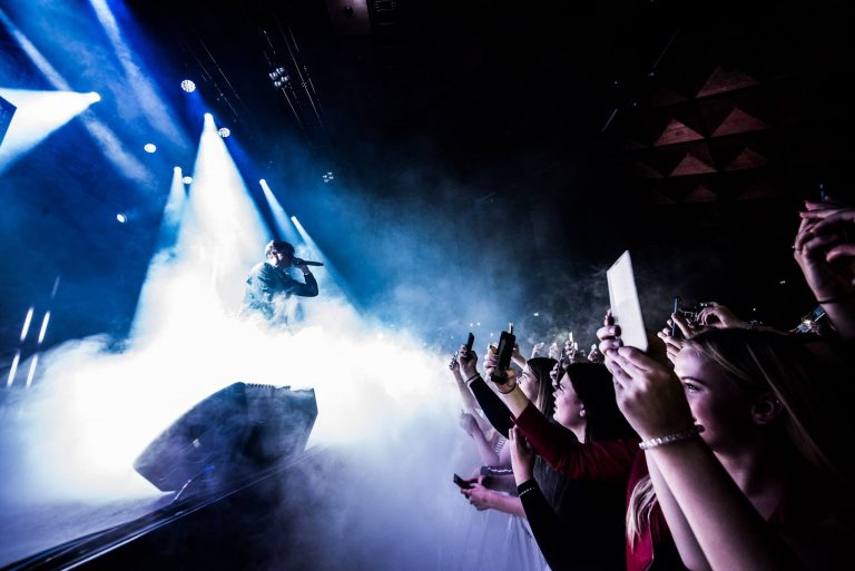 Een artiest performt op een podium omringd door mist, terwijl fans enthousiast hun telefoons omhoog houden.