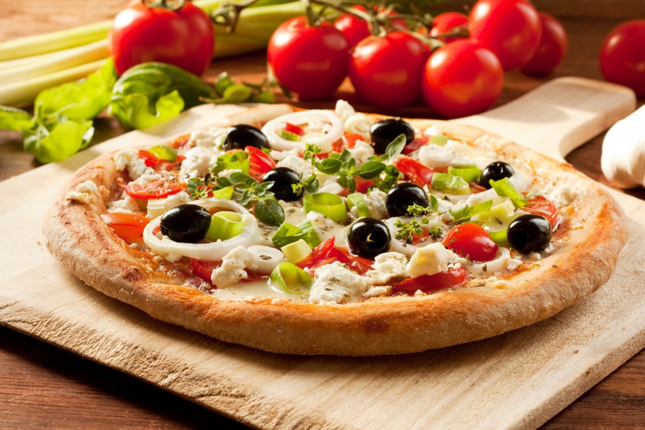 Een versgebakken pizza bedekt met kleurrijke groenten en olijven, omringd door verse tomaten.