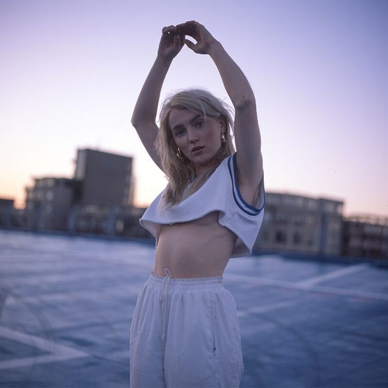 Een vrouw in een sportoutfit staat op een dak bij schemerlicht en rekt haar armen boven haar hoofd.