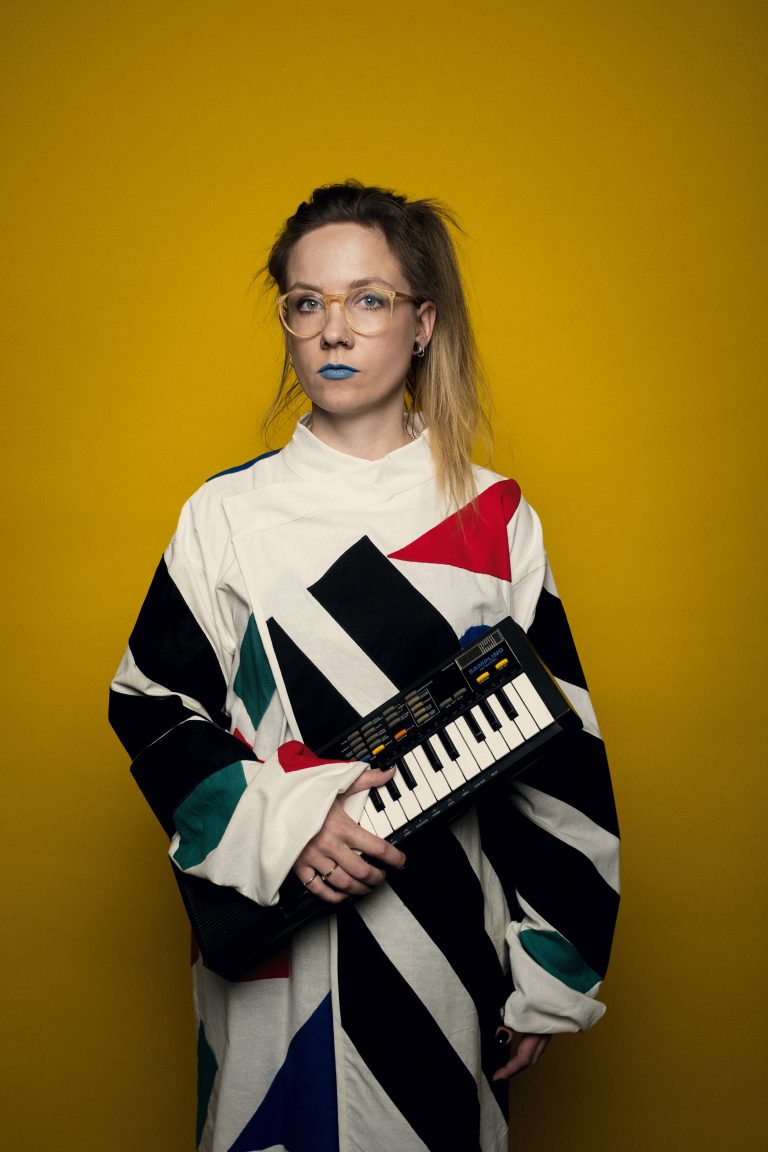 Een persoon met een retro outfit houdt een mini keyboard vast, met een gele achtergrond.