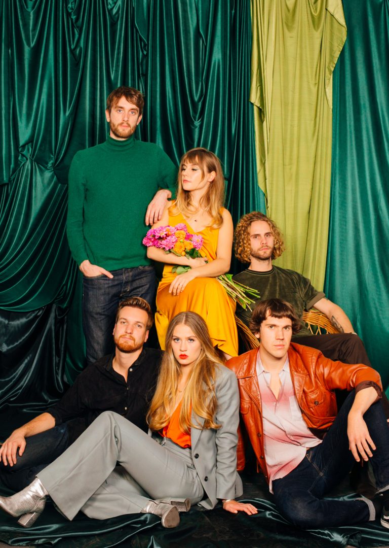 Groep van zes mensen poserende voor een kleurrijke achtergrond met groene en gele stoffen, waarbij een vrouw in een gele jurk bloemen vasthoudt.