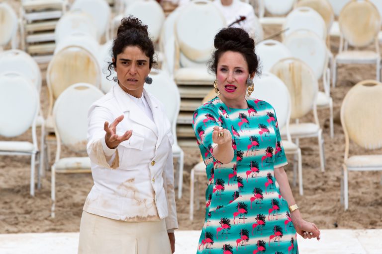 Twee vrouwen met opvallende outfits staan naast elkaar en strekken hun handen uit, met lege stoelen op de achtergrond.