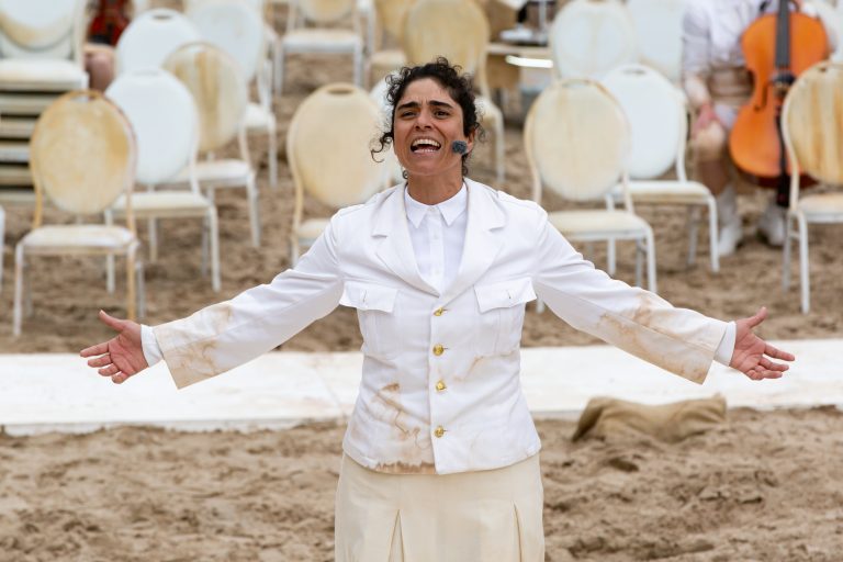 Een vrouw in een witte outfit staat op een strand met uitgestrekte armen en een blije uitdrukking, omringd door lege stoelen.