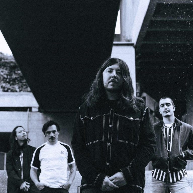 Een zwart-witfoto van vier mannen die onder een viaduct staan, met de man in het midden die een lange, donkere jas draagt.