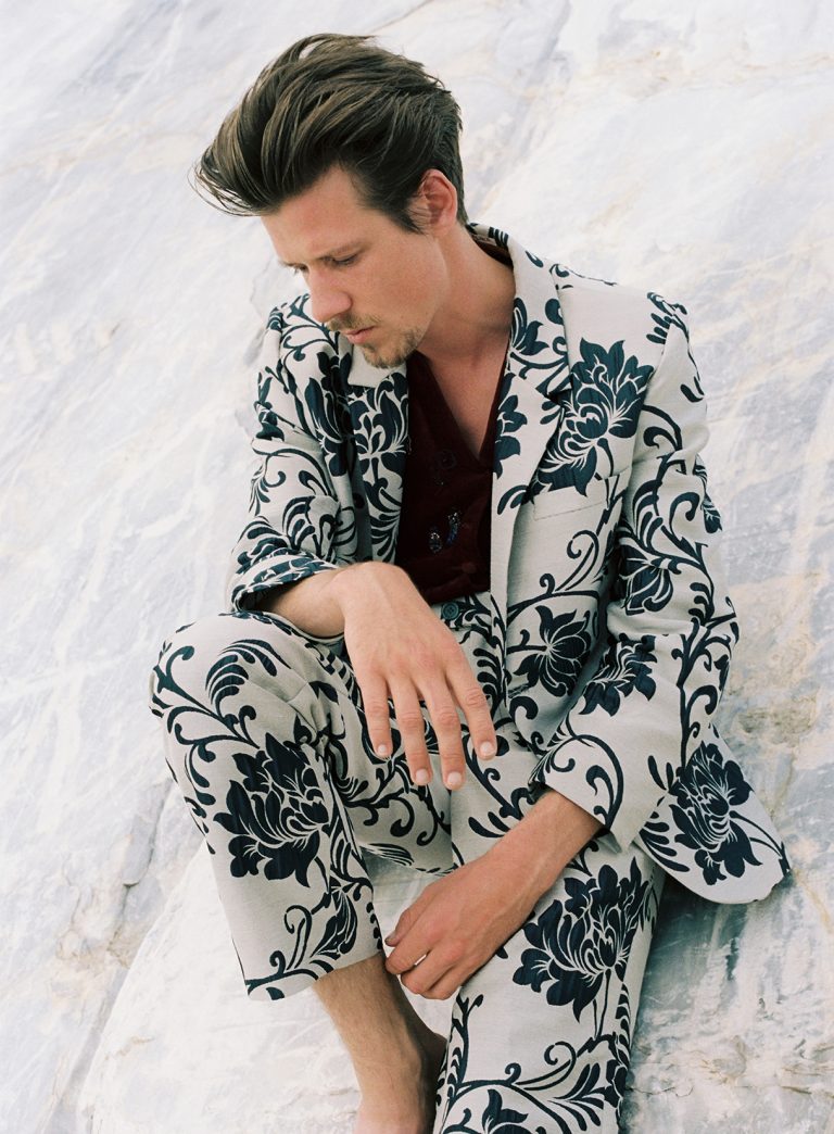 Een man in een opvallend zwart-wit gebloemd pak zit op een lichte ondergrond en kijkt pensive.