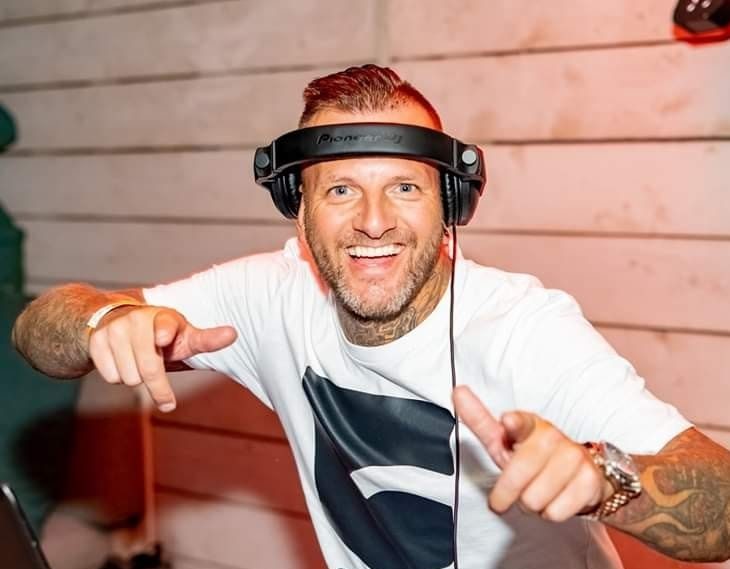 Een glimlachende DJ in een wit shirt met een zwarte print, terwijl hij de handen in de lucht steekt en muziek draait.