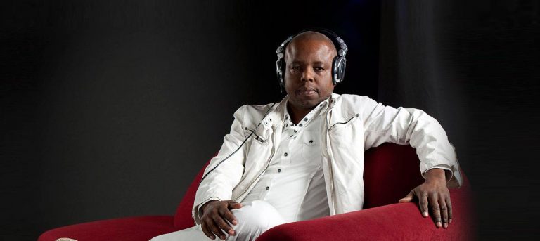 Een man in een witte outfit zit op een rode bank, luisterend naar muziek met hoofdtelefoon op.