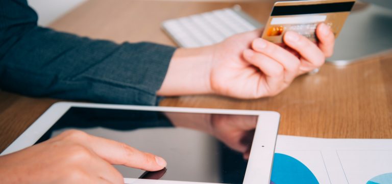 Een hand houdt een creditcard boven een tablet die op een bureau ligt, terwijl de andere hand de tablet aanraakt.