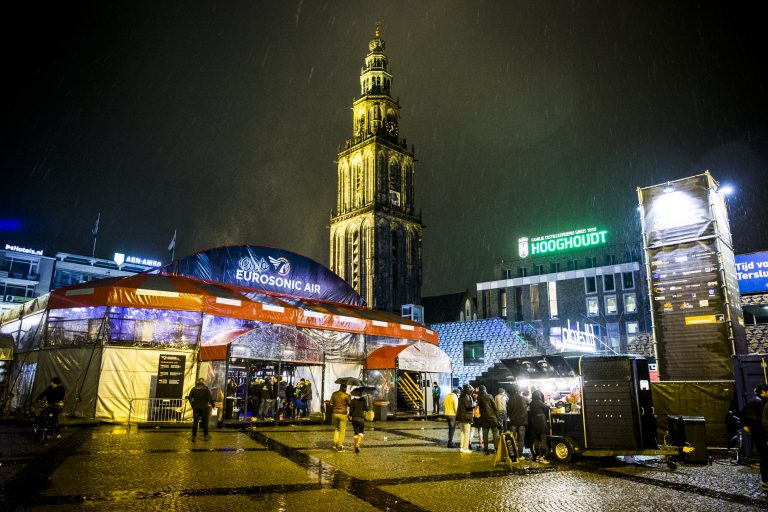 Een stadsplein met een toren op de achtergrond, verlicht door nachtverlichting en omgeven door bezoekers en kraampjes.