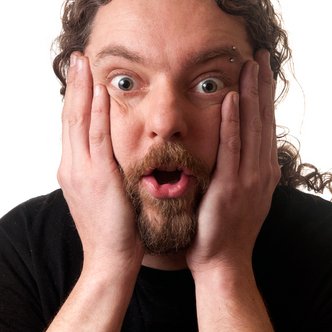 Een man met lang, krullend haar, die geschokt kijkt en zijn handen op zijn wangen houdt.