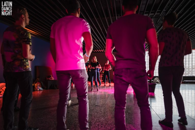 Twee mensen staan van achteren gezien en kijken naar een groep dansers in een fel verlichte ruimte.
