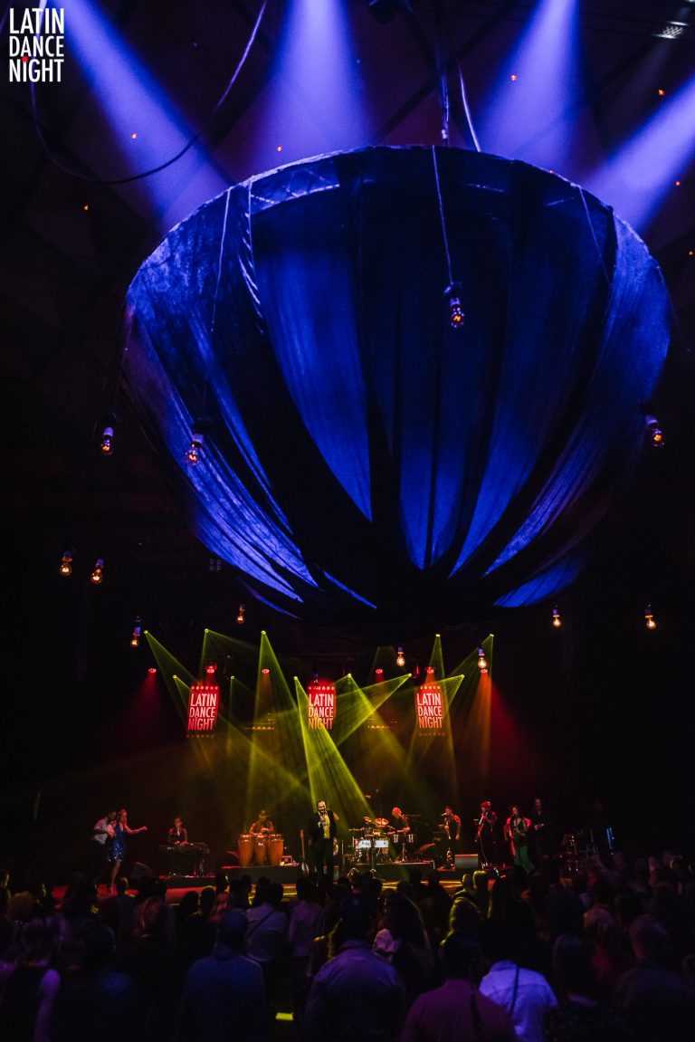 Zicht op een podium met een band, verlicht door kleurrijke lichten en een grote, blauwe doek aan het plafond.