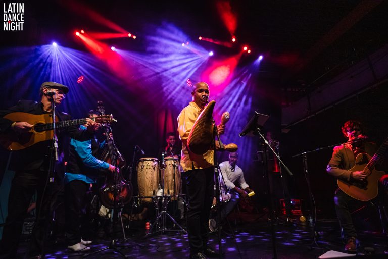 Een muzikant in een gele shirt speelt op een handpan, omringd door andere bandleden onder sfeervolle verlichting.