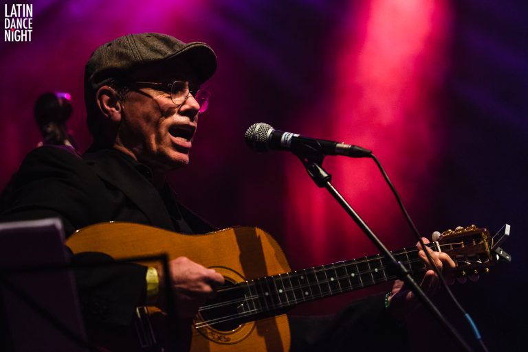 Een muzikant zingt met een gitaar terwijl kleurrijke lichten op de achtergrond schijnen.