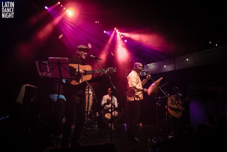 Een live band speelt muziek op een podium, verlicht door kleurrijke lichten.