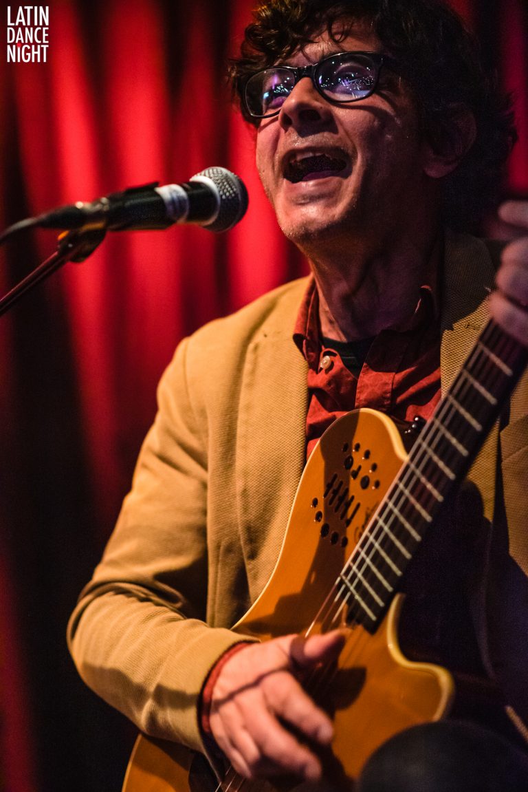 Een muzikant zingt en speelt gitaar op een podium met een rode achtergrond.