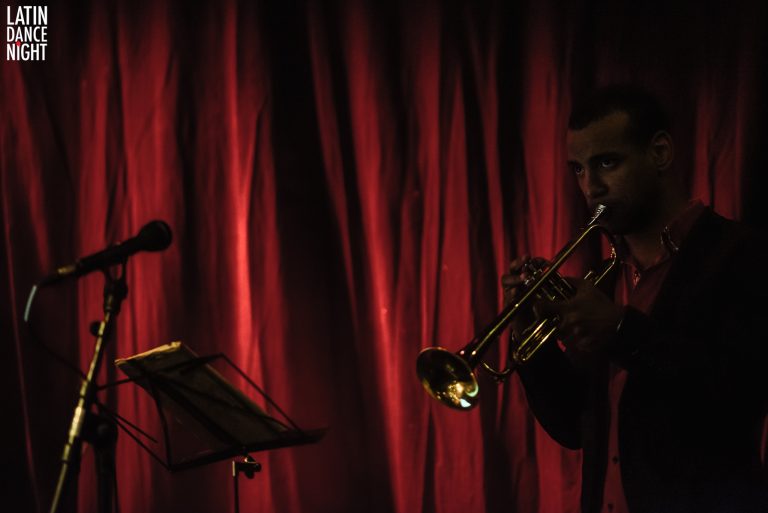 Een muzikant speelt trompet op een podium met een rode achtergrond.