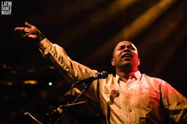 Een zanger met uitgestoken arm staat achter een microfoon en is zichtbaar in een levendige muzikale uitvoering.