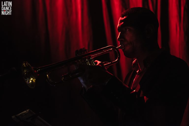 Een muzikant speelt trompet in een sfeervolle, gedimde omgeving met rode verlichting.