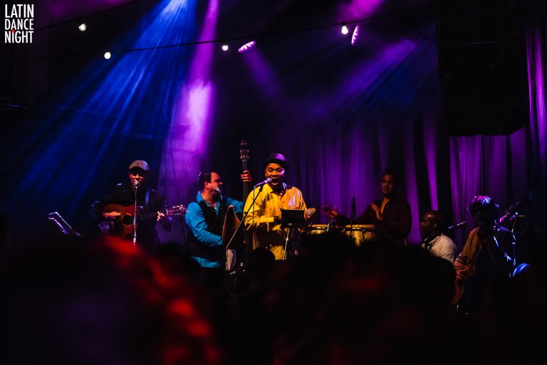 Een muzikale groep speelt op een podium onder paarse en blauwe verlichting.