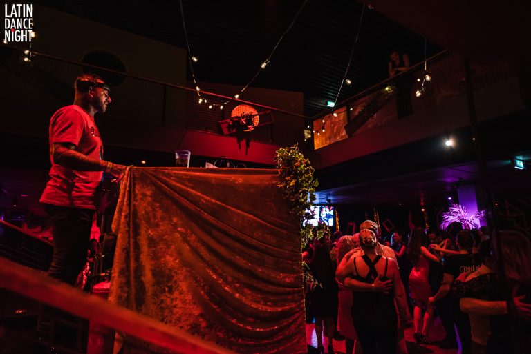 Een vertoning van een levendige avond met mensen in een sfeervolle ruimte, gedimd licht en een podium in de hoogte.