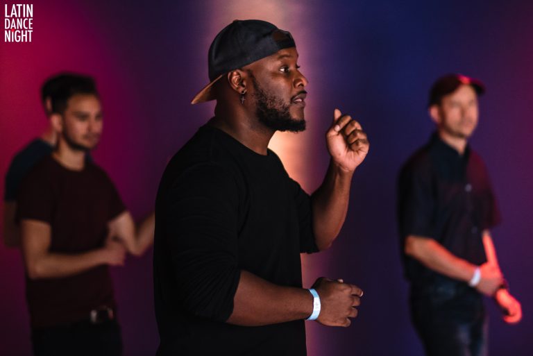 Een man in een zwart T-shirt en pet houdt een spreekbeurt terwijl anderen op de achtergrond staan.