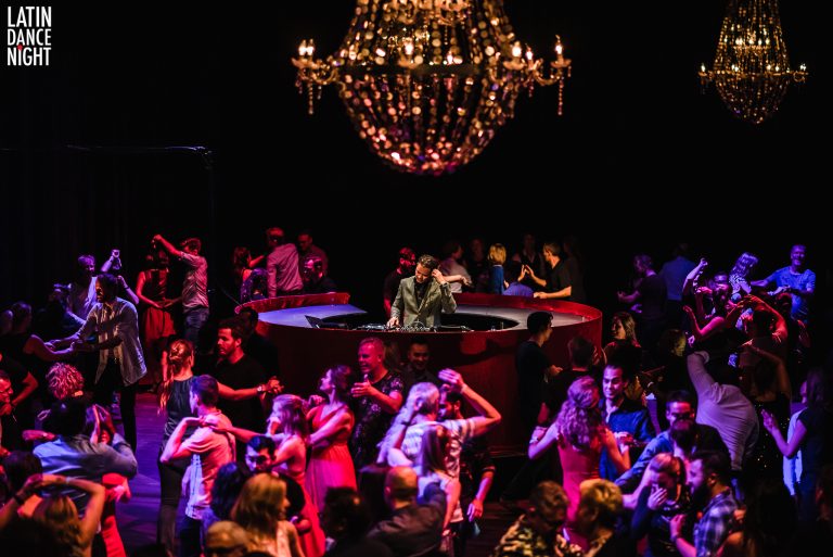 Een drukke dansvloer met mensen die dansen onder een grote kroonluchter, verlicht in sfeervolle kleuren.