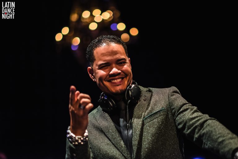 Een glimlachende DJ in een glanzend groen jasje, met een headset en een feestelijke achtergrond.