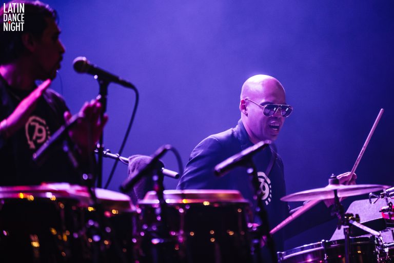 Een drummer speelt op een podium met een paar conga's onder een blauwe verlichting.