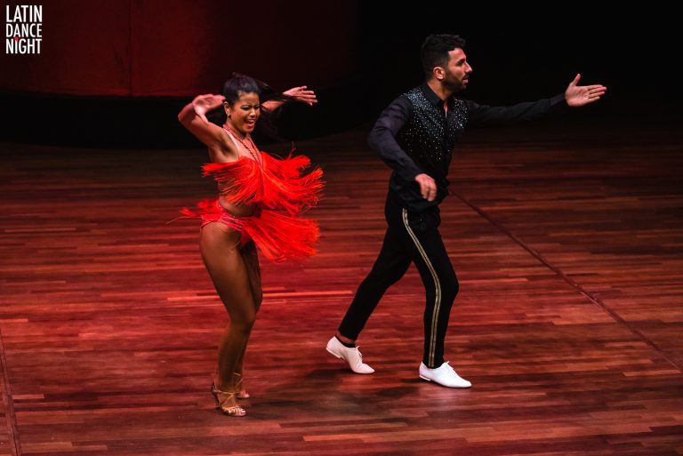Een man en een vrouw dansen op een houten podium, de vrouw in een levendige rode dansoutfit met een uitlopende rok.