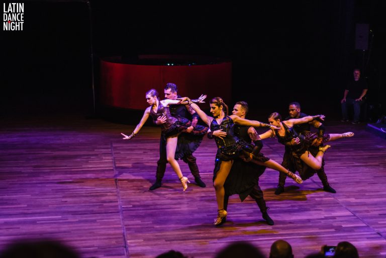 Een groep dansers in zwarte outfits voert een dynamische choreografie uit op een podium met paarse verlichting.