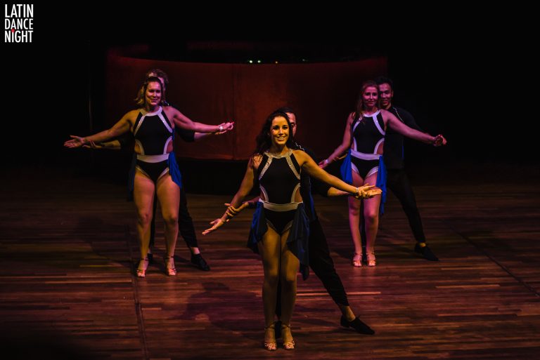 Een groep dansers voert een choreografie uit op een podium, gekleed in sportieve outfits.
