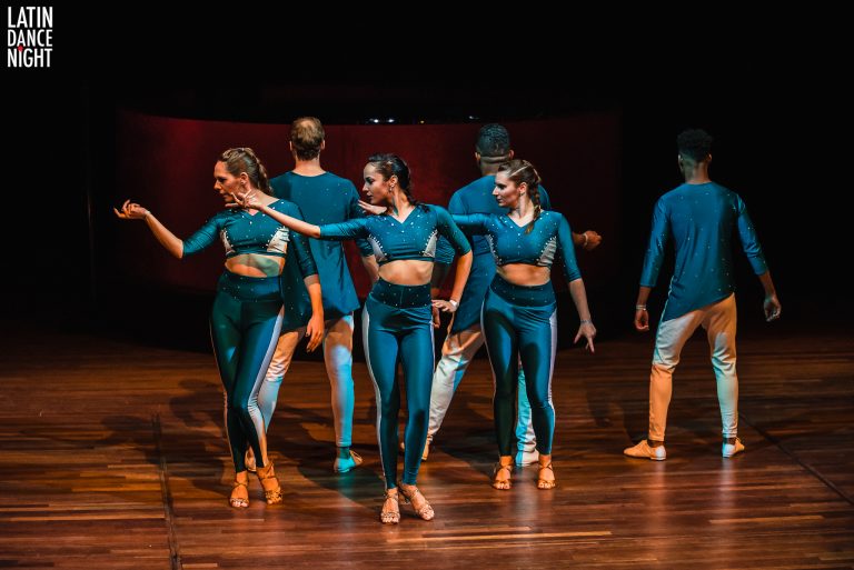 Groep dansers in blauwe outfits voert een choreografie uit op een houten podium.